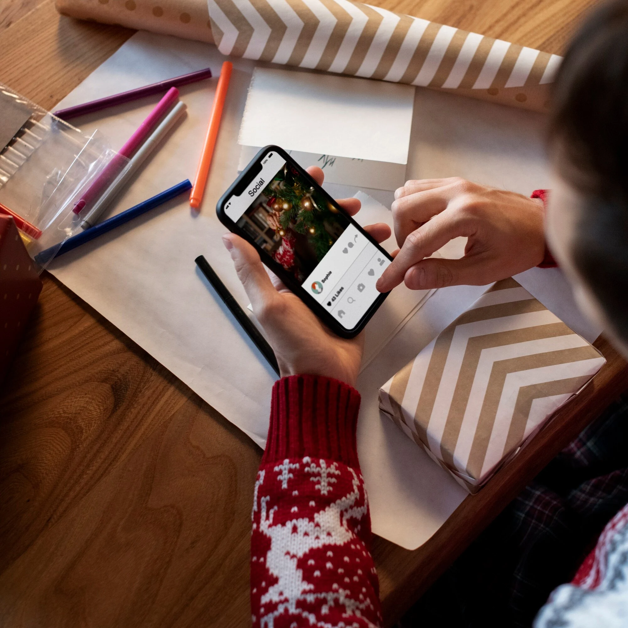 Person in a festive sweater using a smartphone at a wooden table, surrounded by gift wrap, colored pens, and a wrapped present, creating a cozy holiday atmosphere.